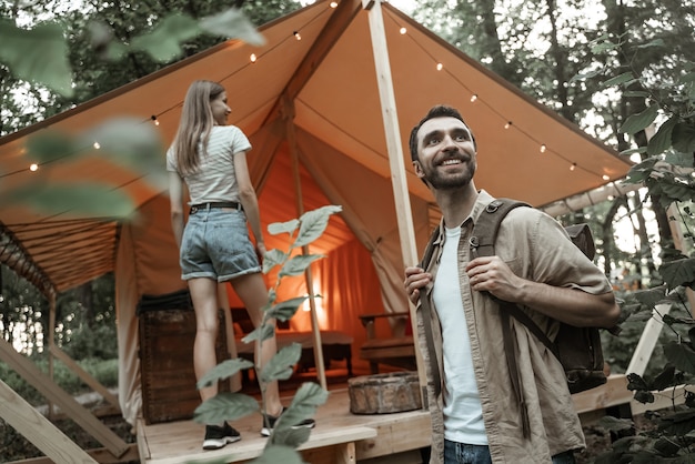 Pareja romántica acampando al aire libre y de pie en la tienda glamping. Feliz el hombre y la mujer en unas románticas vacaciones de camping. Hombre con mochila con su novia millennials llegó al sitio de glamping.