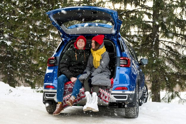 Pareja romántica joven inconformista abrazándose mientras está sentado en el maletero del coche bajo la nieve que cae