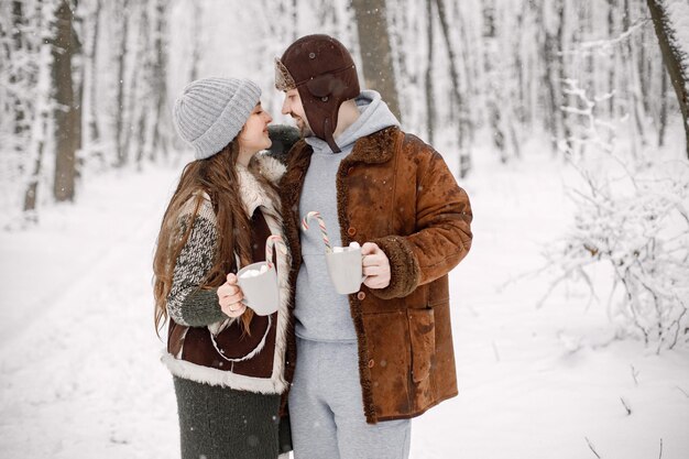 Pareja romántica de pie en el bosque de invierno abrazando y sosteniendo dos tazas con bebida de cacao
