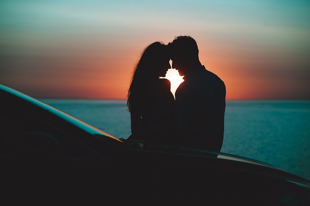 La pareja romántica de pie cerca del coche en el hermoso fondo del atardecer