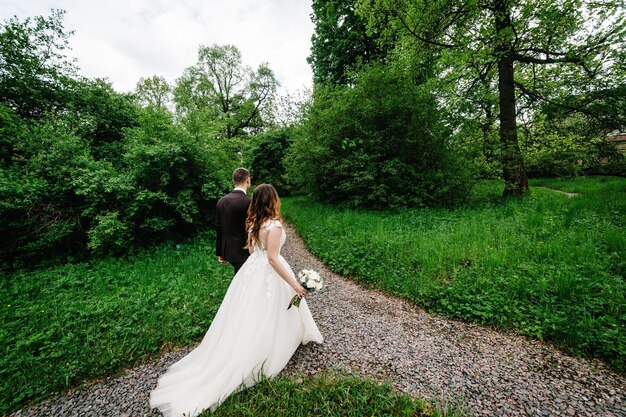 Pareja romántica recién casados novia y novio está caminando de regreso por un sendero en un parque verde Momento de boda feliz y alegre