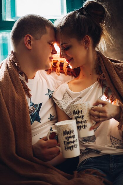 Pareja romántica sentada en la colina de las ventanas con luces con una taza creativa en la mano tocándose por las cabezas Novio y novia vestidos con camiseta blanca y jeans abrazándose y disfrutando del amor