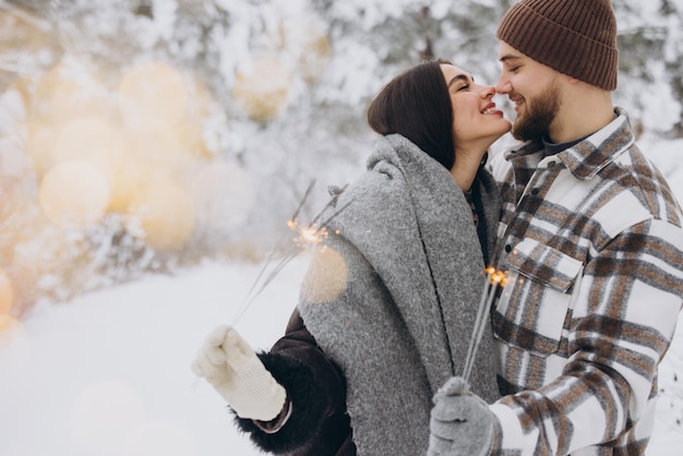 Foto una pareja romántica sonriente con sombreros de punto posando con chispas en el bosque de invierno sosteniendo luces de bengala