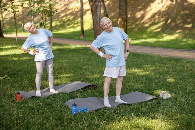 Pareja senior alegre en ropa deportiva hace curvas laterales en el parque de la ciudad