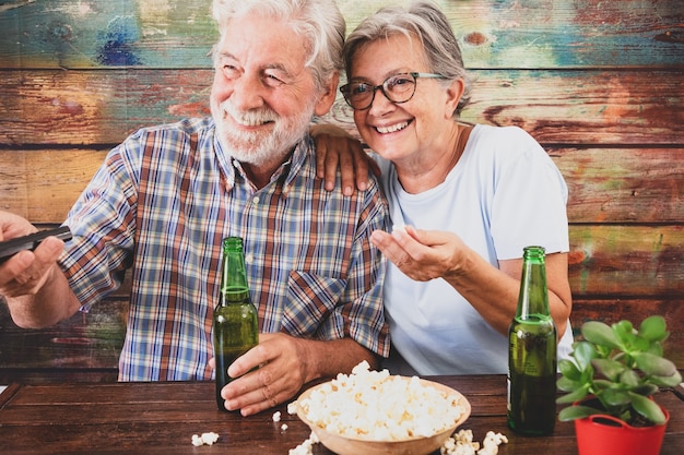 Pareja senior alegre viendo un partido de fútbol en la televisión, bebiendo cerveza y comiendo palomitas de maíz. Fondo de madera