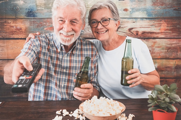 Pareja senior alegre viendo un partido de fútbol en la televisión, bebiendo cerveza y comiendo palomitas de maíz. Fondo de madera