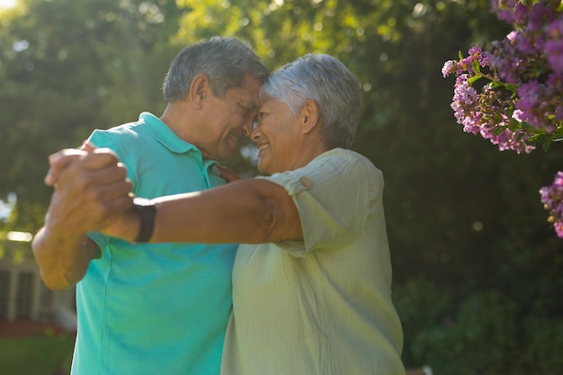 Pareja senior birracial romántica sonriente mirándose mientras bailan contra árboles en el parque