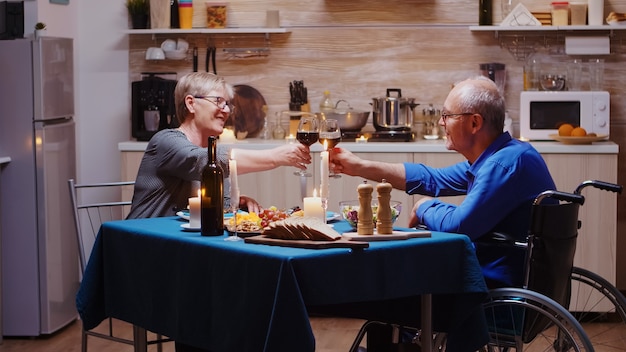 Pareja senior brindando copas sentados a la mesa en la cocina. Marido en silla de ruedas discapacitados paralizados inmovilizados con una cena romántica con su esposa alegre