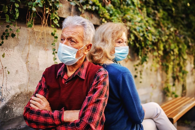 Pareja senior enojada con máscaras protectoras al sentarse en el banco espalda con espalda.