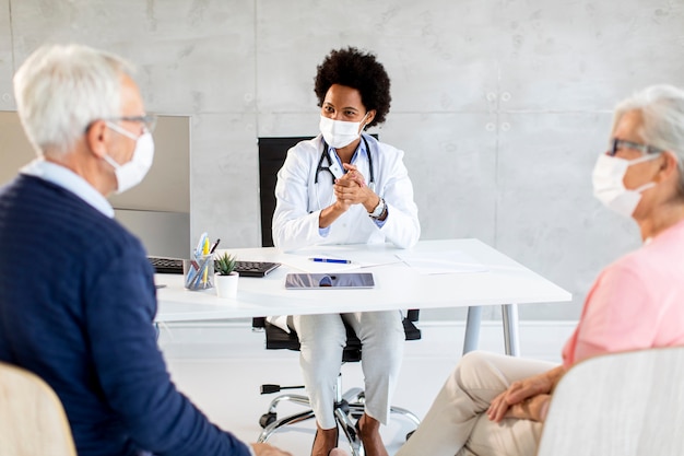 Pareja senior con máscaras faciales protectoras recibe noticias de doctora negra en la oficina