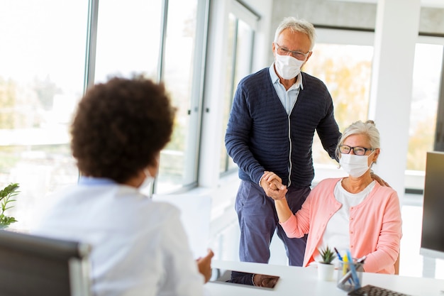 Pareja senior con máscaras faciales protectoras recibe noticias de doctora negra en la oficina