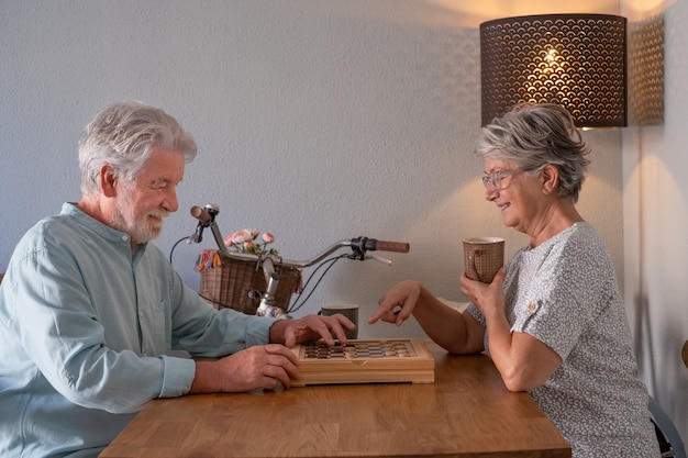 Pareja senior relajada pasar tiempo juntos en casa jugando un juego de damas en la mesa de madera.