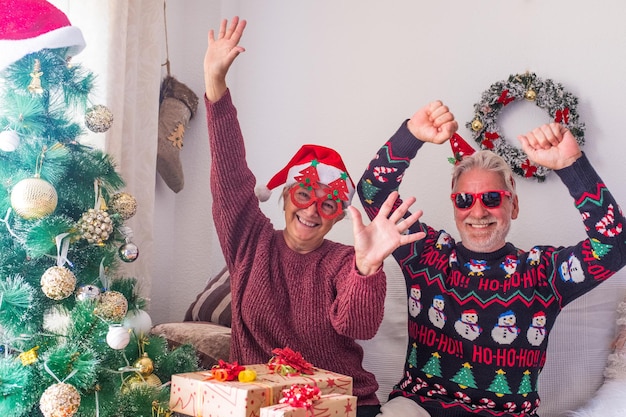 Pareja senior en ropa de abrigo y gorro de Papá Noel bailando y celebrando frente al árbol de Navidad decorado en casa. Amantes de la vieja pareja heterosexual romántica celebrando juntos el festival de navidad