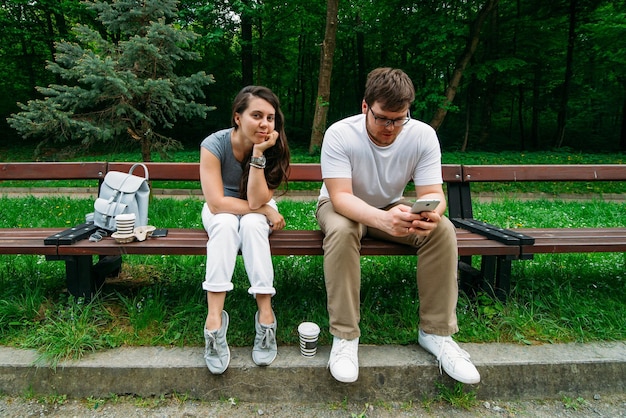 Pareja sentada en un banco en el parque de la ciudad, la mujer es un hombre aburrido mirando el teléfono inteligente