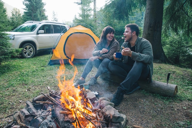 Pareja sentada cerca del fuego del campamento y bebiendo té y contando historias carpa y suv en el fondo