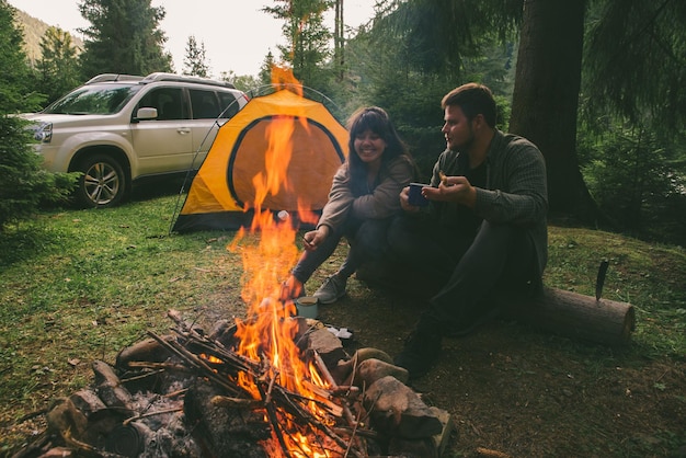 Pareja sentada cerca del fuego del campamento y bebiendo té y contando historias carpa y suv en el fondo