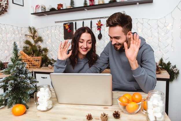 Una pareja sentada en la cocina usa una computadora portátil con una llamada de videochat hablando con familiares amigos agitando las manos