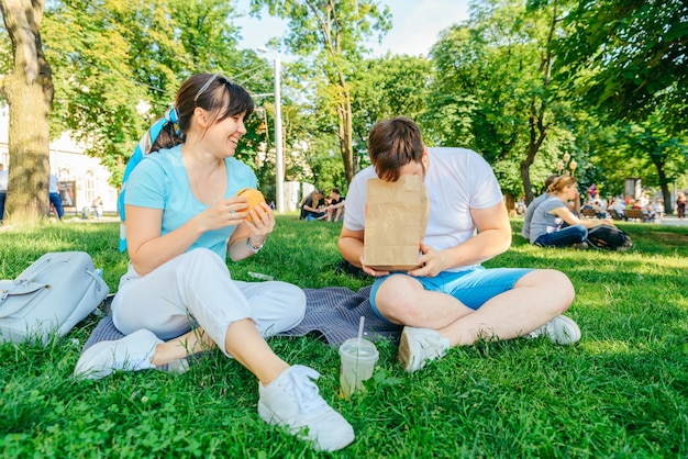 Pareja Sentada en la hierba verde comiendo hamburguesas bebiendo batido angel ancho