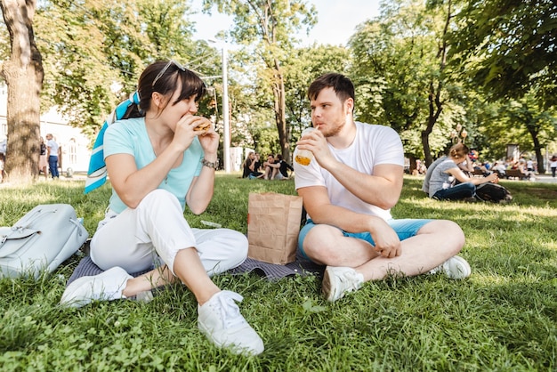 Pareja Sentada en la hierba verde comiendo hamburguesas bebiendo batido