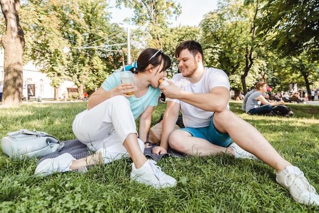 Pareja Sentada en la hierba verde comiendo hamburguesas bebiendo batido