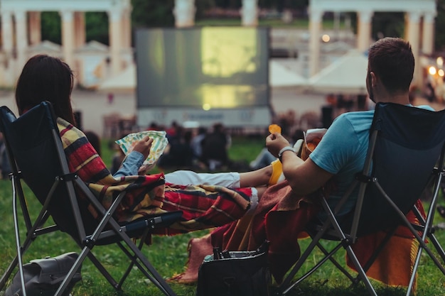 Pareja sentada en sillas de camping en el parque de la ciudad mirando películas al aire libre en el cine al aire libre