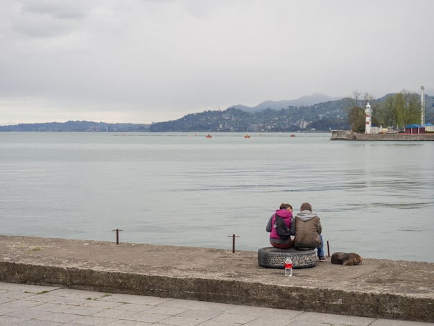 Una pareja sentada sobre un neumático en el puerto junto a un perro Silencio y paz