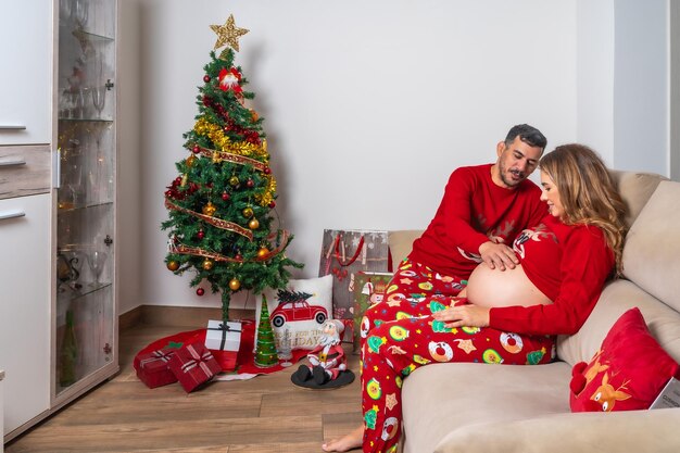 Pareja sentada en el sofá junto al árbol de Navidad abrazando el barrido Familia con mujer embarazada