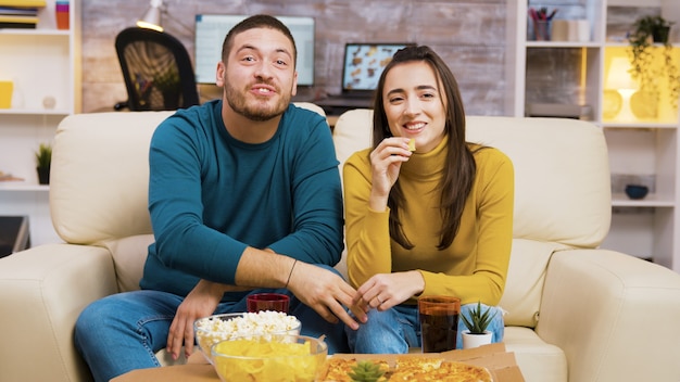 Pareja sentada en el sofá riendo mientras ve la televisión y come pizza. Palomitas de maíz y pizza en la mesa de café.