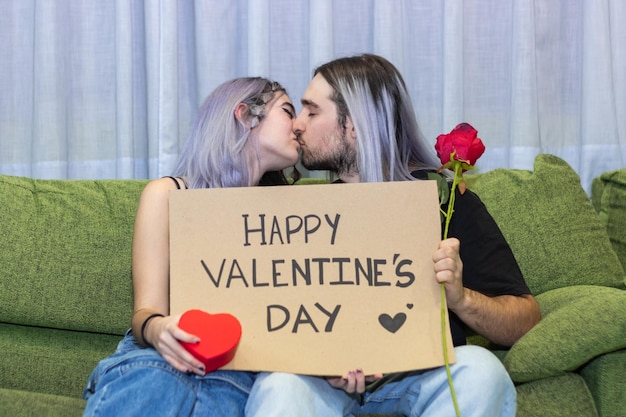 Pareja sentada en el sofá sosteniendo un cartel con el texto del Día de San Valentín Pareja joven besándose sentada en el sofá sosteniendo un cartel del Día de San Valentín