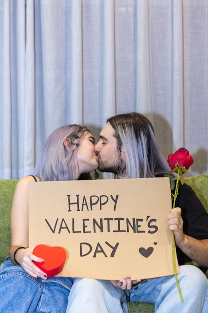 Pareja sentada en el sofá sosteniendo un cartel con el texto del Día de San Valentín Pareja joven besándose sentada en el sofá sosteniendo un cartel del Día de San Valentín