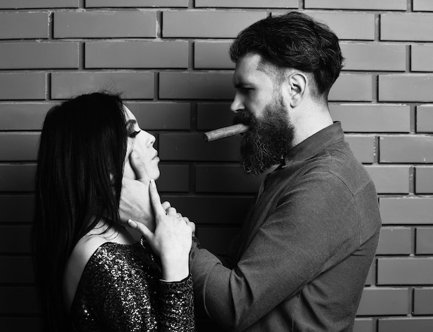 Pareja seria de hombre barbudo barba larga brutal hipster caucásico con bigote y mujer bonita o chica sexy con maquillaje de cabello moreno en vestido de lentejuelas verdes sostiene cigarro en la pared de ladrillo