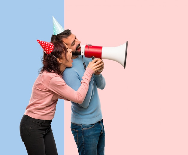 Pareja con sombreros de cumpleaños y sosteniendo un megáfono sobre fondo rosa y azul