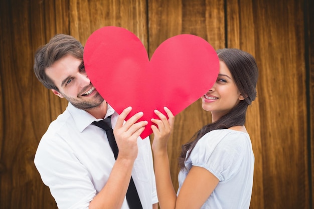 Pareja sonriendo a la cámara sosteniendo un corazón contra la mesa de madera