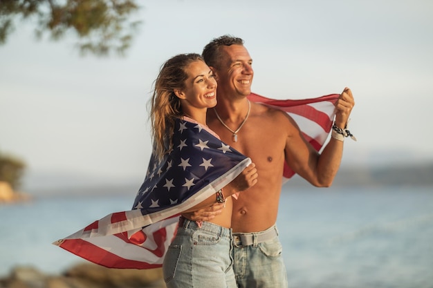 Una pareja sonriente con bandera nacional americana disfrutando de un relajante día en la playa.