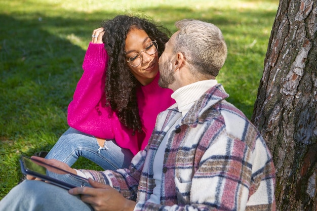 Una pareja sonriente y feliz sentada en el césped del parque mirándose a los ojos usando tecnología moderna