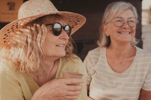 Una pareja sonriente y optimista de mujeres mayores y de mediana edad con sombreros hacen confidencias beben una taza de café