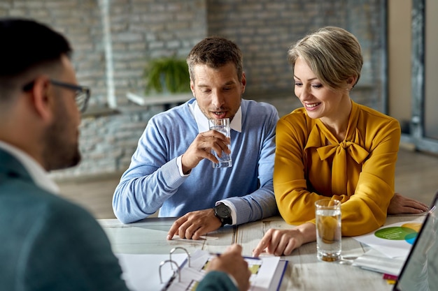 Pareja sonriente y su agente inmobiliario analizando planos en una reunión en la oficina