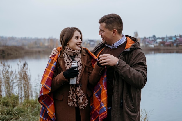 Foto pareja sonriente tomando una bebida caliente junto al lago con manta