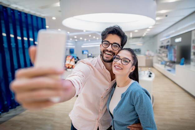 Pareja sonriente tomando selfie mientras está de pie en la tienda de tecnología. Concepto de tecnologías contemporáneas.