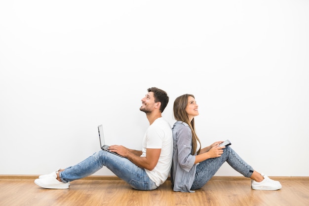 La pareja de la sonrisa se sienta con una computadora portátil y una tableta cerca de la pared vacía