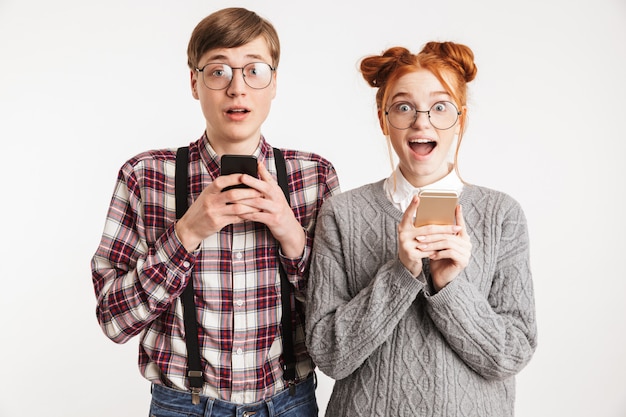 Pareja sorprendida de nerds escolares usando teléfonos móviles