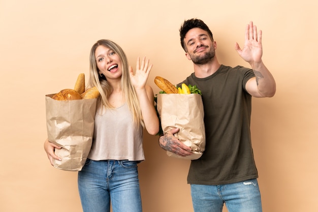 Pareja sosteniendo bolsas de la compra de comestibles sobre saludando con la mano con expresión feliz