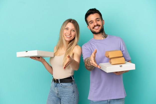 Pareja sosteniendo pizzas y hamburguesas sobre fondo azul aislado un apretón de manos para cerrar un buen trato