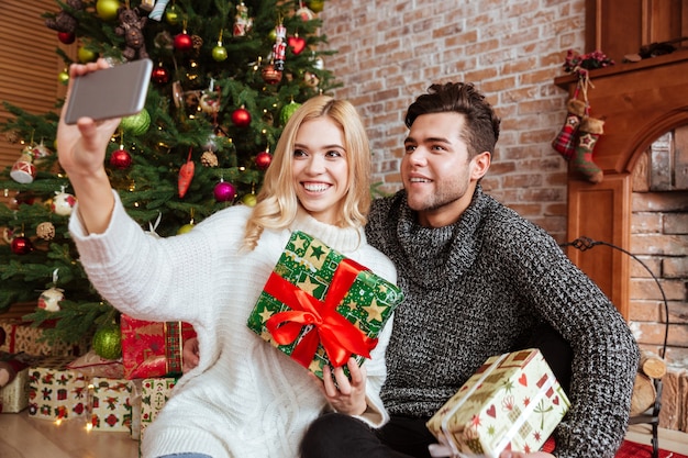 Pareja en suéteres sentada en el suelo y hace selfie con regalo