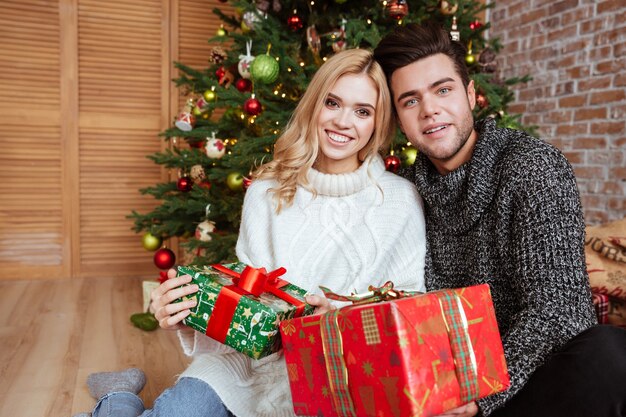 Pareja en suéteres sentados en el suelo con regalos cerca del abeto