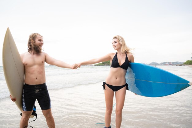 Una pareja de surfistas tomados de la mano y mirándose el uno al otro en la playa