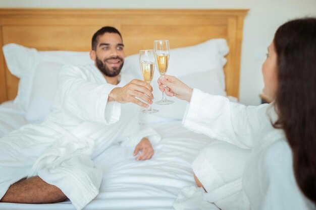 Pareja tintineo de vasos bebiendo vino espumoso vistiendo albornoces en el hotel