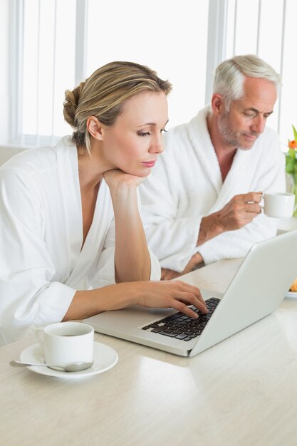 Pareja tomando un café en el desayuno en batas de baño usando la computadora portátil