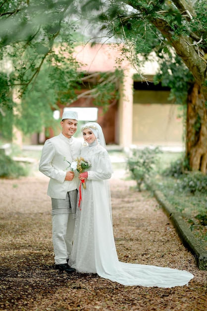 Una pareja con traje tradicional de boda se para en un parque.