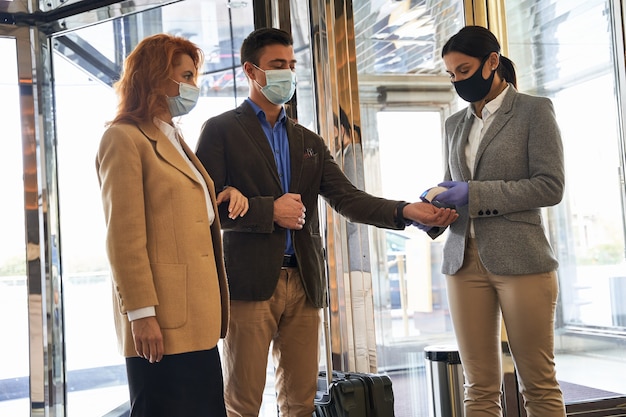 Pareja de turistas con máscaras médicas entrando en el pasillo del hotel y una mujer controlando su temperatura